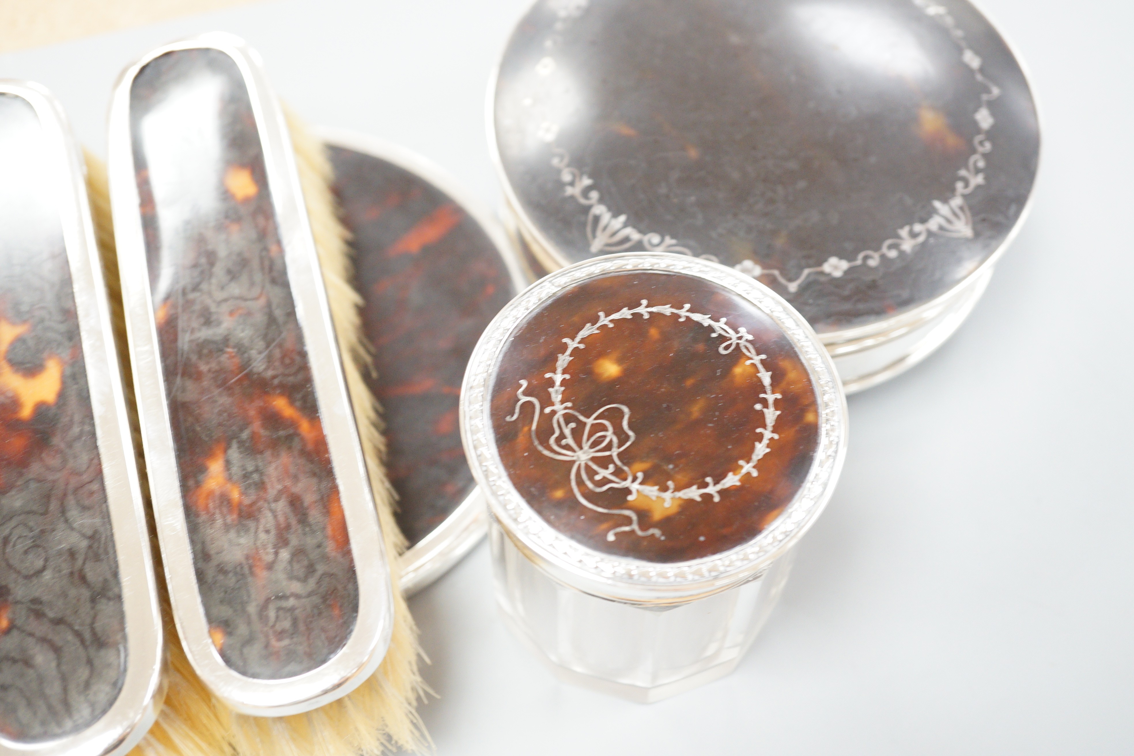A George V matched silver and tortoiseshell mounted five piece mirror and brush set, a George V silver and tortoiseshell pique mounted circular box with mirrored interior and a similar mounted glass toilet jar.